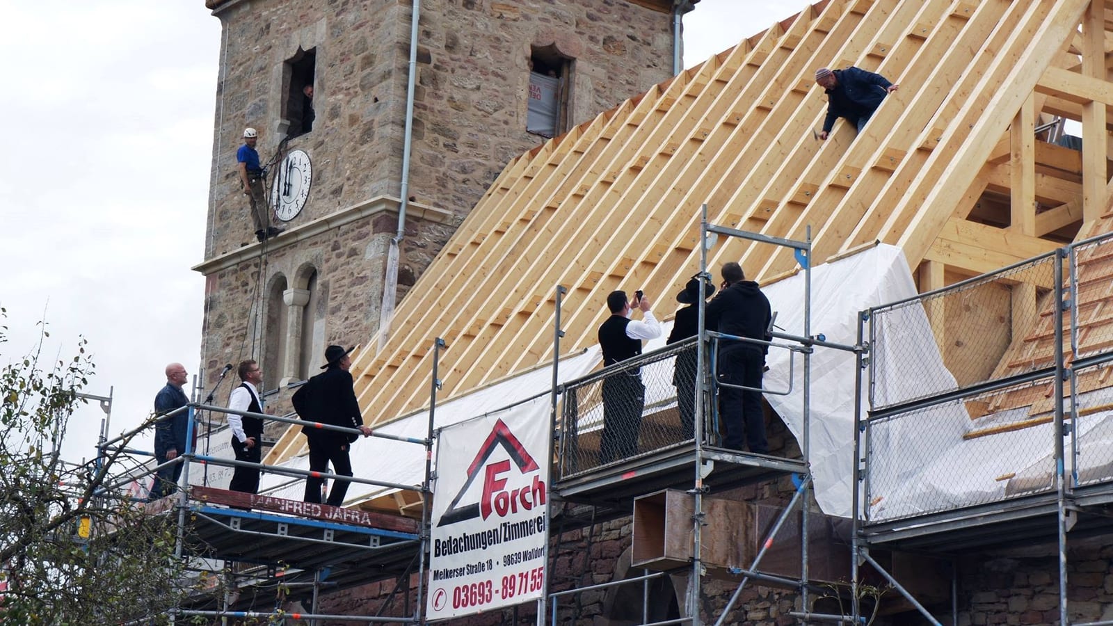 Gäste und Handwerker auf dem Gerüst vor dem Kirchturm der Kirchenburg Walldorf bei Meiningen, daneben der neue Dachstuhl des Kirchenschiffs und ein Forch-Banner am Gerüst